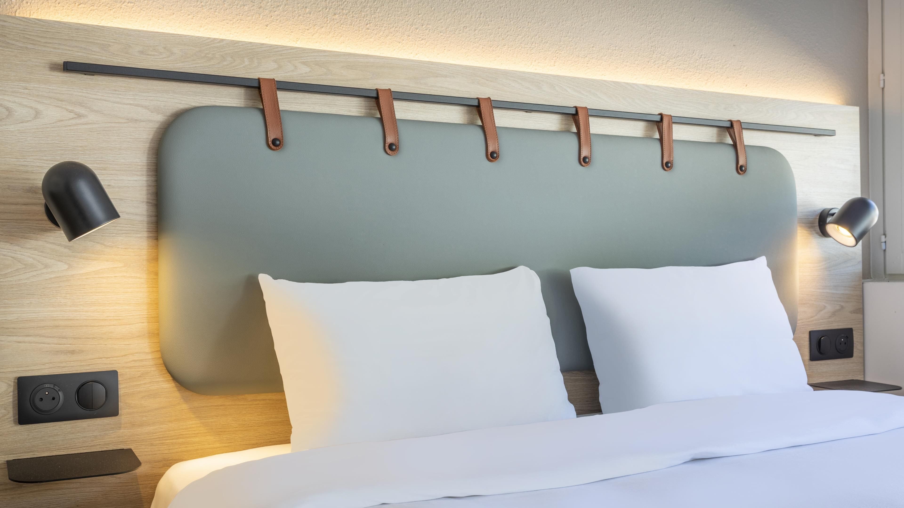 A modern hotel room with a large bed, two bedside lamps, a wooden headboard with leather straps, and a wall-mounted control panel.