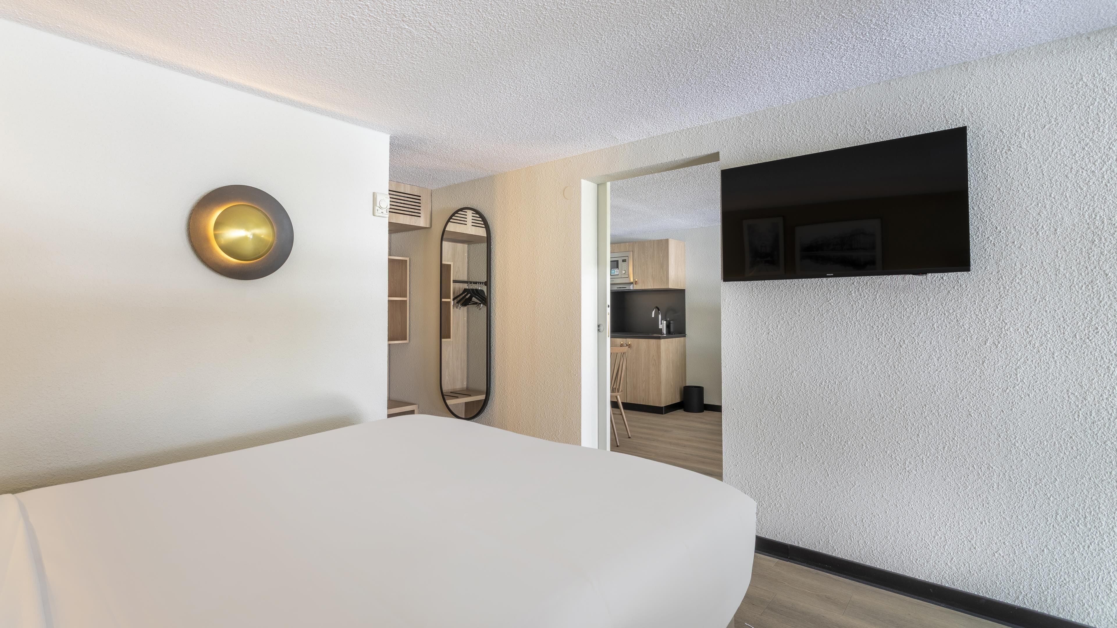 Ein modernes Hotelzimmer mit einem großen Bett, einer Wandleuchte, einem Spiegel und Blick in den Küchenbereich.