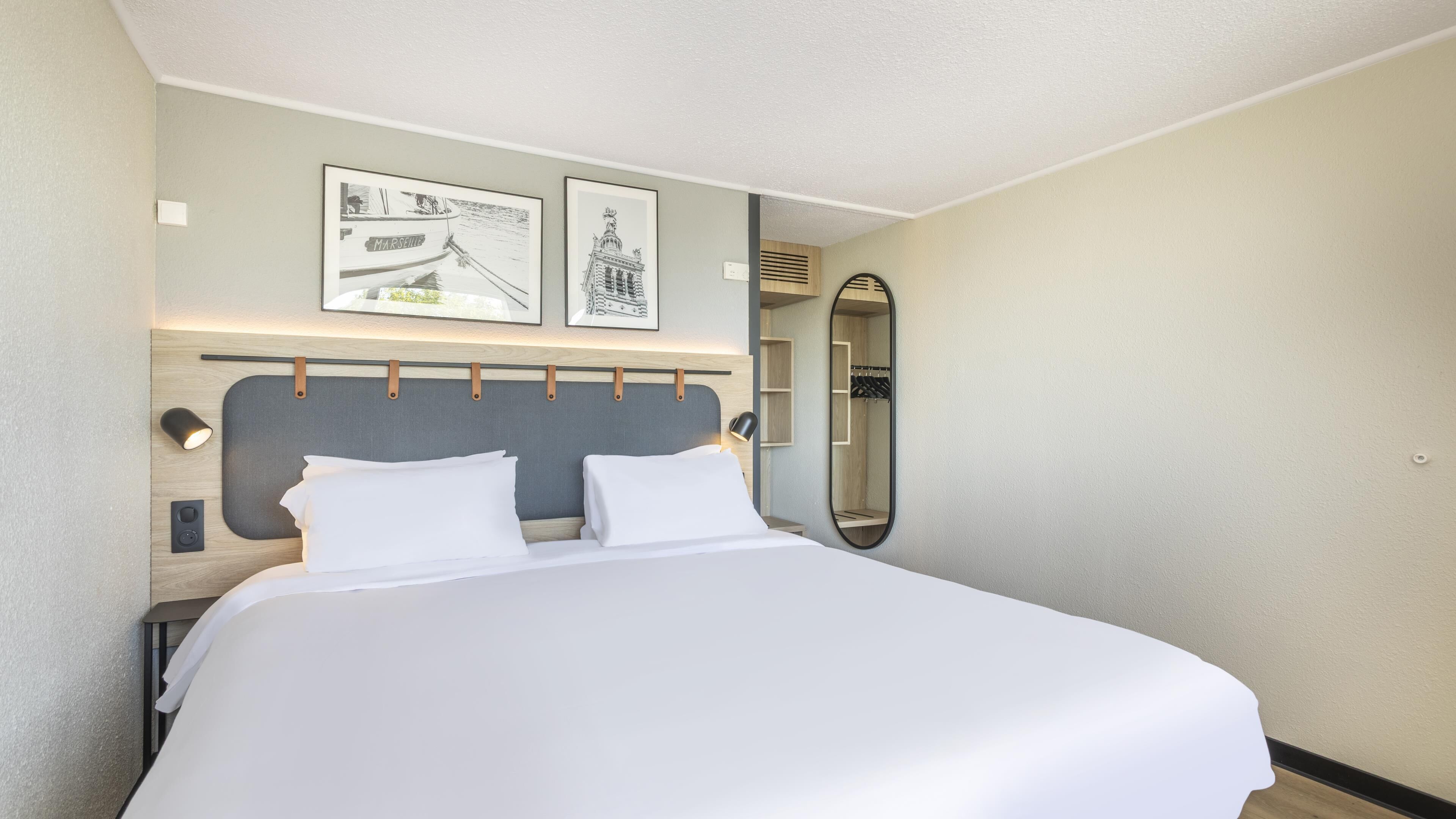 Een net opgemaakt bed in een hotelkamer, met een hoofdbord, twee ingelijste foto's aan de muur, een bedlampje en een grote spiegel.