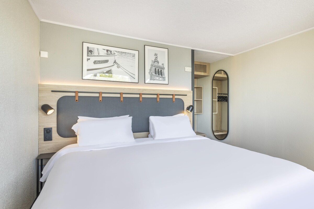 Un lit bien fait dans une chambre d’hôtel, avec une tête de lit, deux tableaux encadrés au mur, une lampe de chevet et un grand miroir.