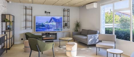 A modern living room with a flat-screen TV, comfortable seating, a fireplace, and large windows offering a view of greenery.