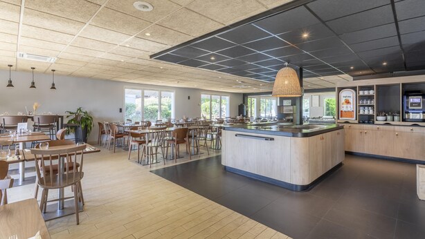 Un restaurante espacioso con una isla central, mesas y sillas de madera y un diseño de techo moderno.