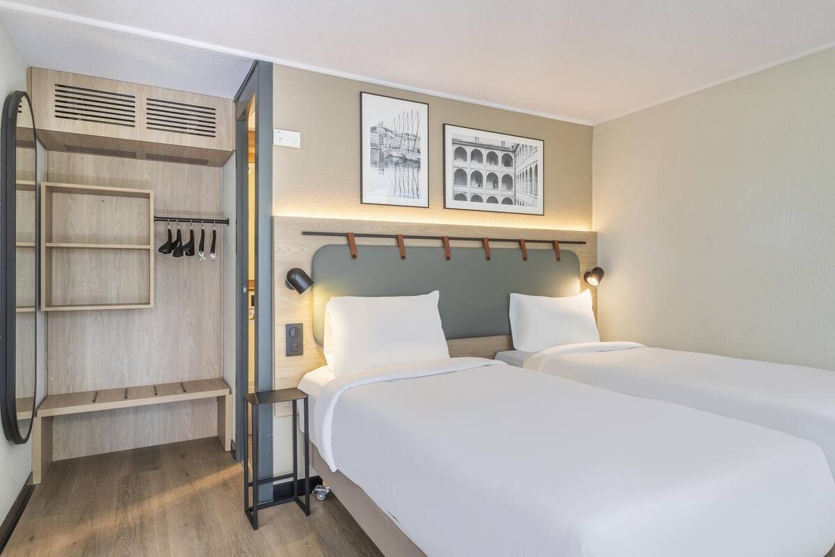 A hotel room with two beds, a wooden wardrobe, and two framed pictures on the wall.