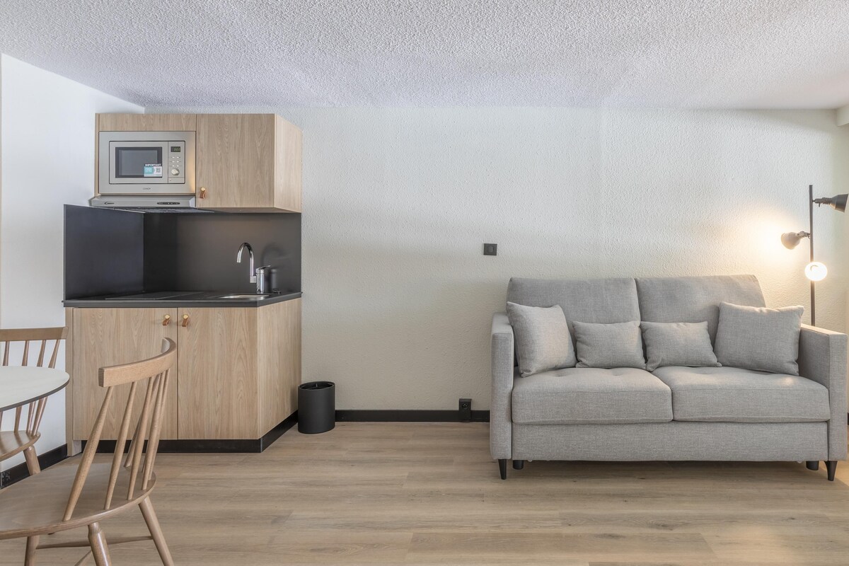 Une cuisine moderne équipée d’un four à micro-ondes encastré, d’un évier et d’un meuble en bois. Le salon comprend un canapé gris et une petite table avec des chaises. Une poubelle noire est visible près du canapé.