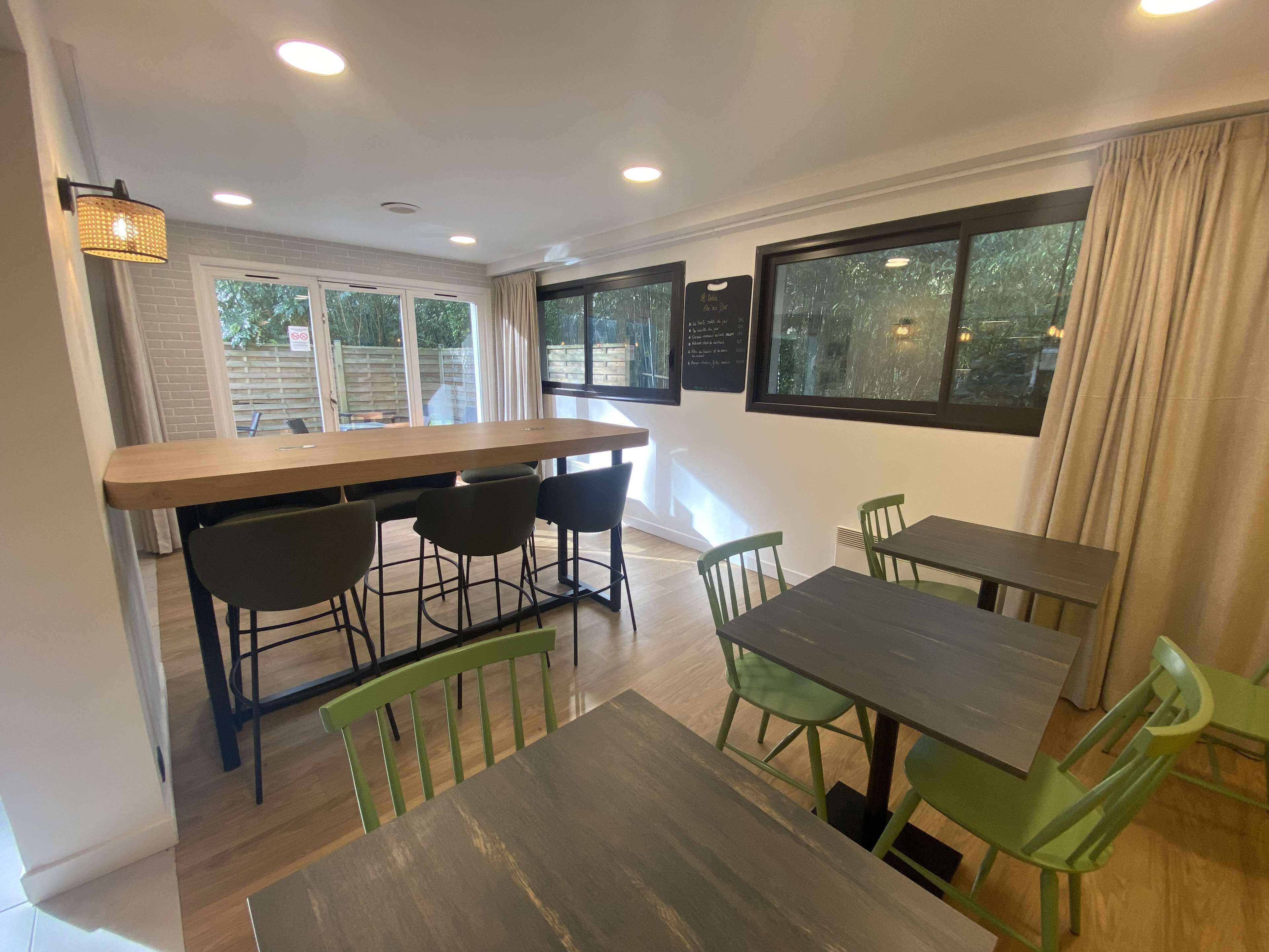 A dining area with a long wooden table and several chairs, large windows with curtains, and a wall-mounted menu board.