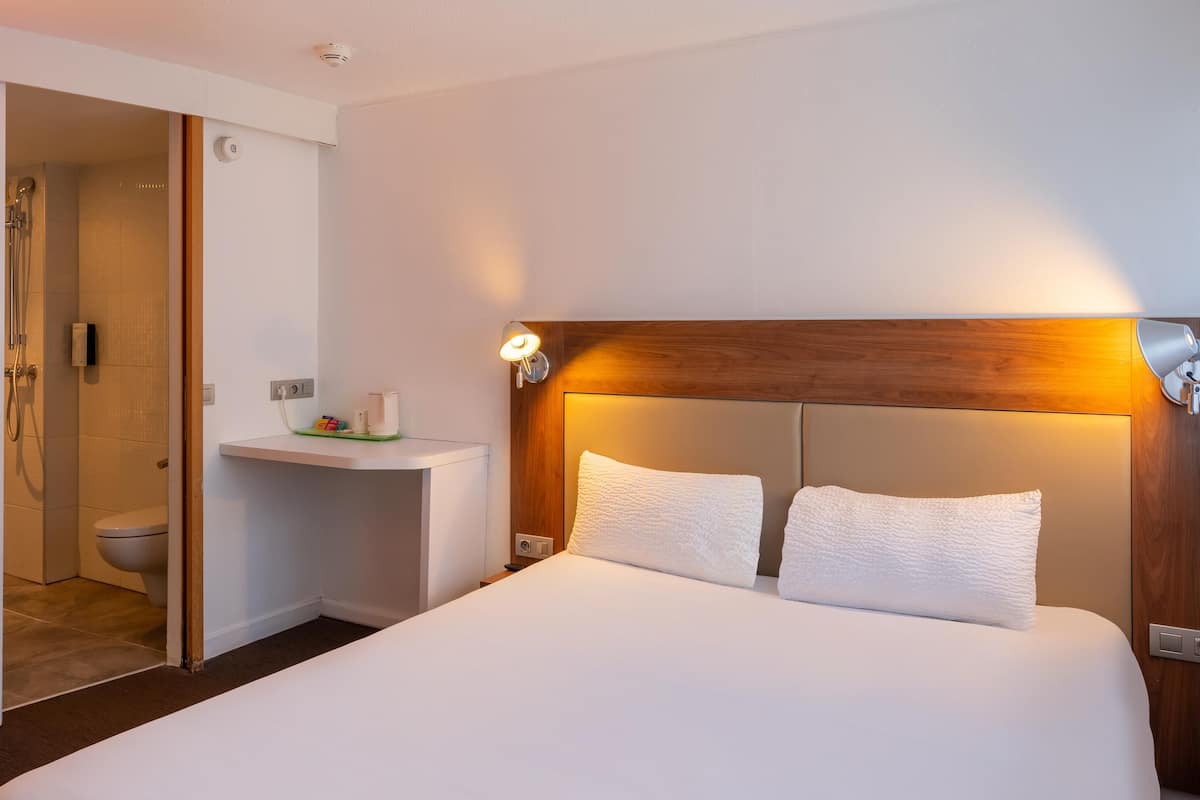 A hotel room with a large bed, a wooden headboard, a bedside table with a lamp, and a shower area.