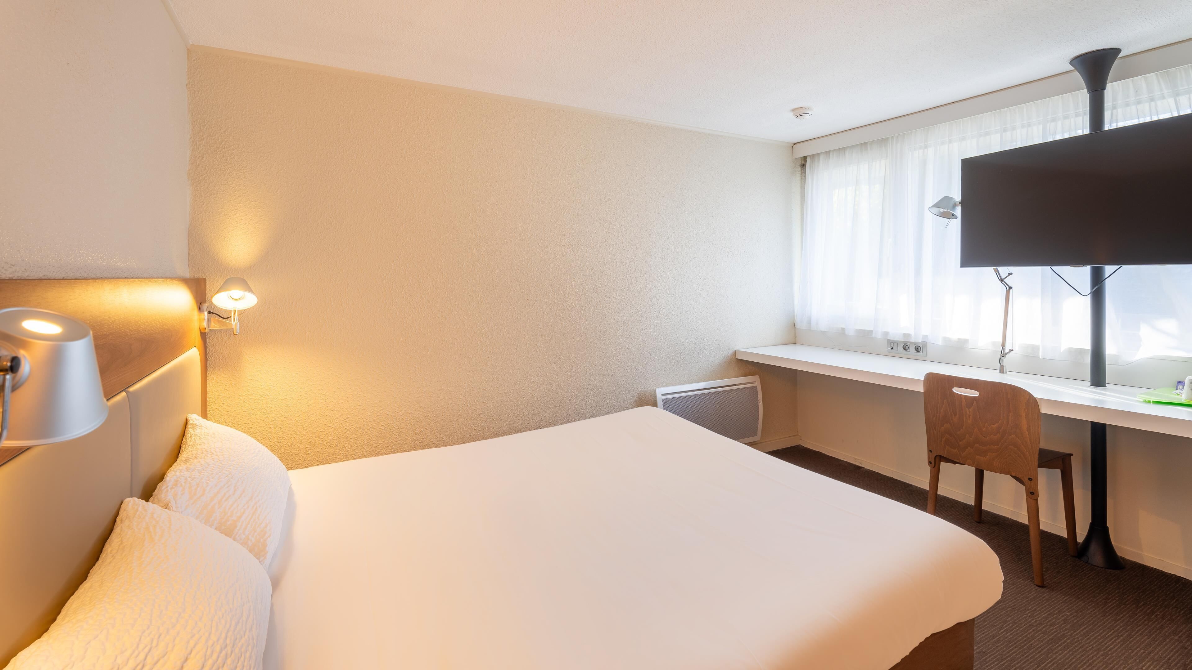 Habitación de hotel con una cama grande, un escritorio, una silla, una lámpara y una ventana con cortinas.