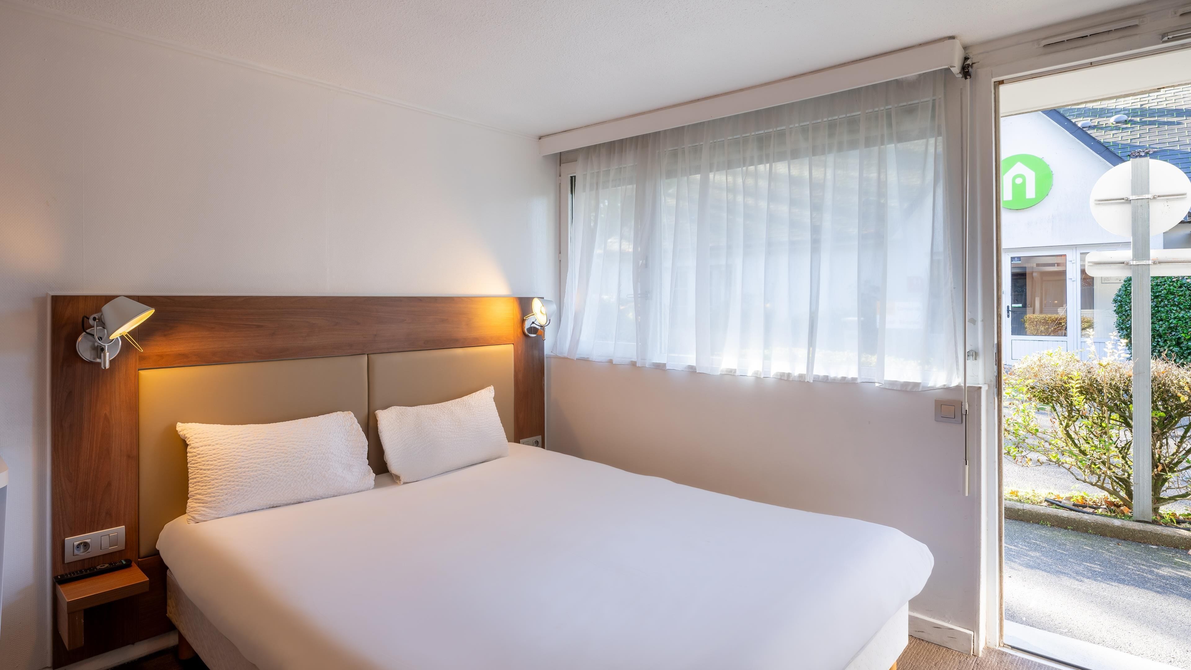 A hotel room with a bed, bedside lamps, a window with sheer curtains, and a view of the outside.