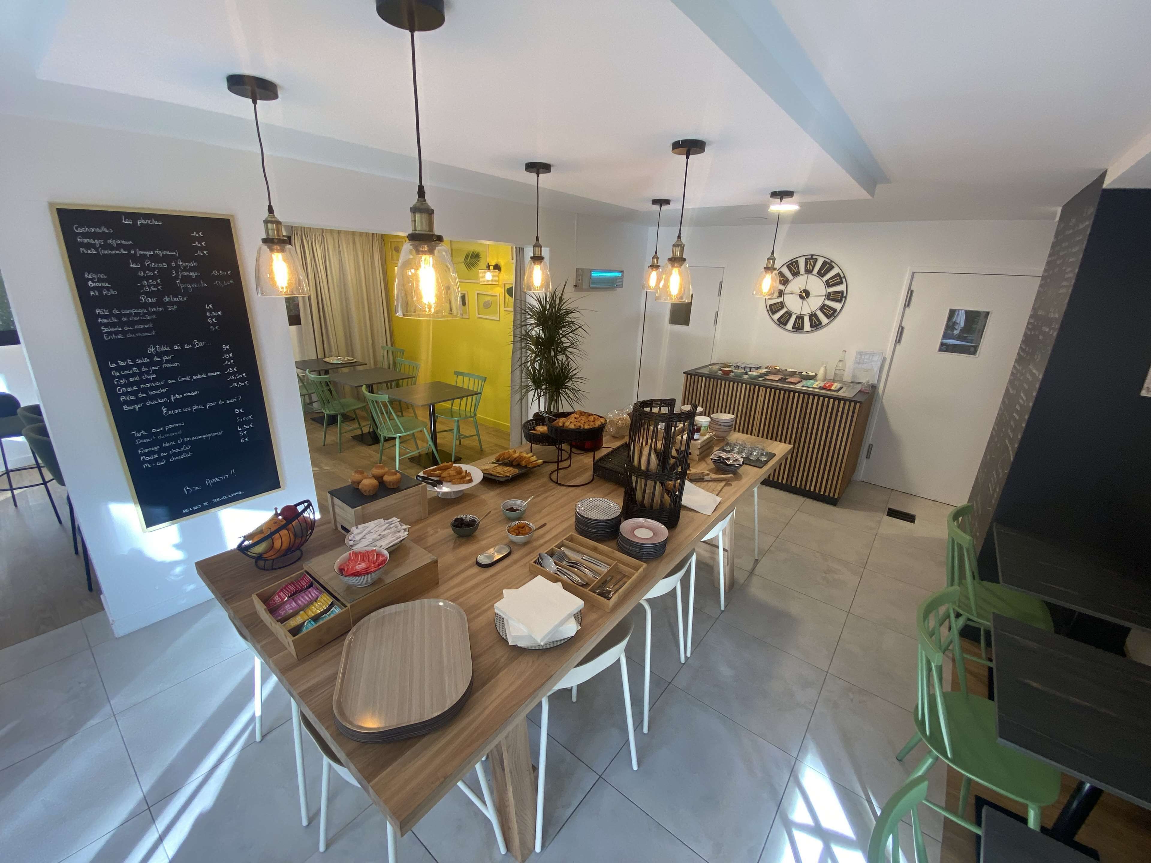 Ein moderner Essbereich mit einem Holztisch, weißen Stühlen und hängenden Pendelleuchten. Eine Kreidetafel mit der Speisekarte ist an der Wand sichtbar.
