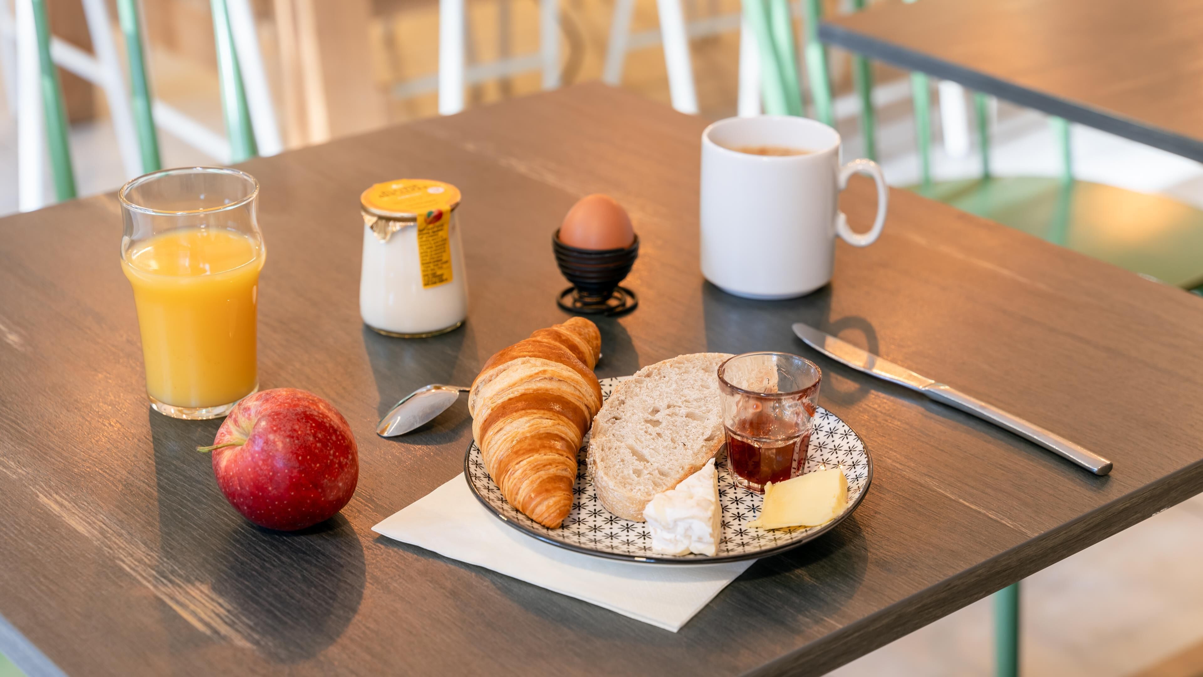 Una mesa de desayuno con cruasanes, un vaso de jugo de naranja, un plato con mantequilla, mermelada y una rebanada de pan, un vaso de té, una jarra de leche y un huevo duro.