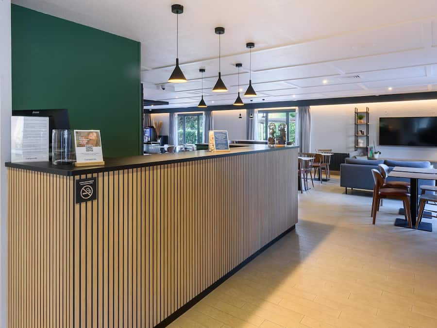 A hotel reception area with a wooden reception desk, a green accent wall, and a seating area with a sofa and dining table.