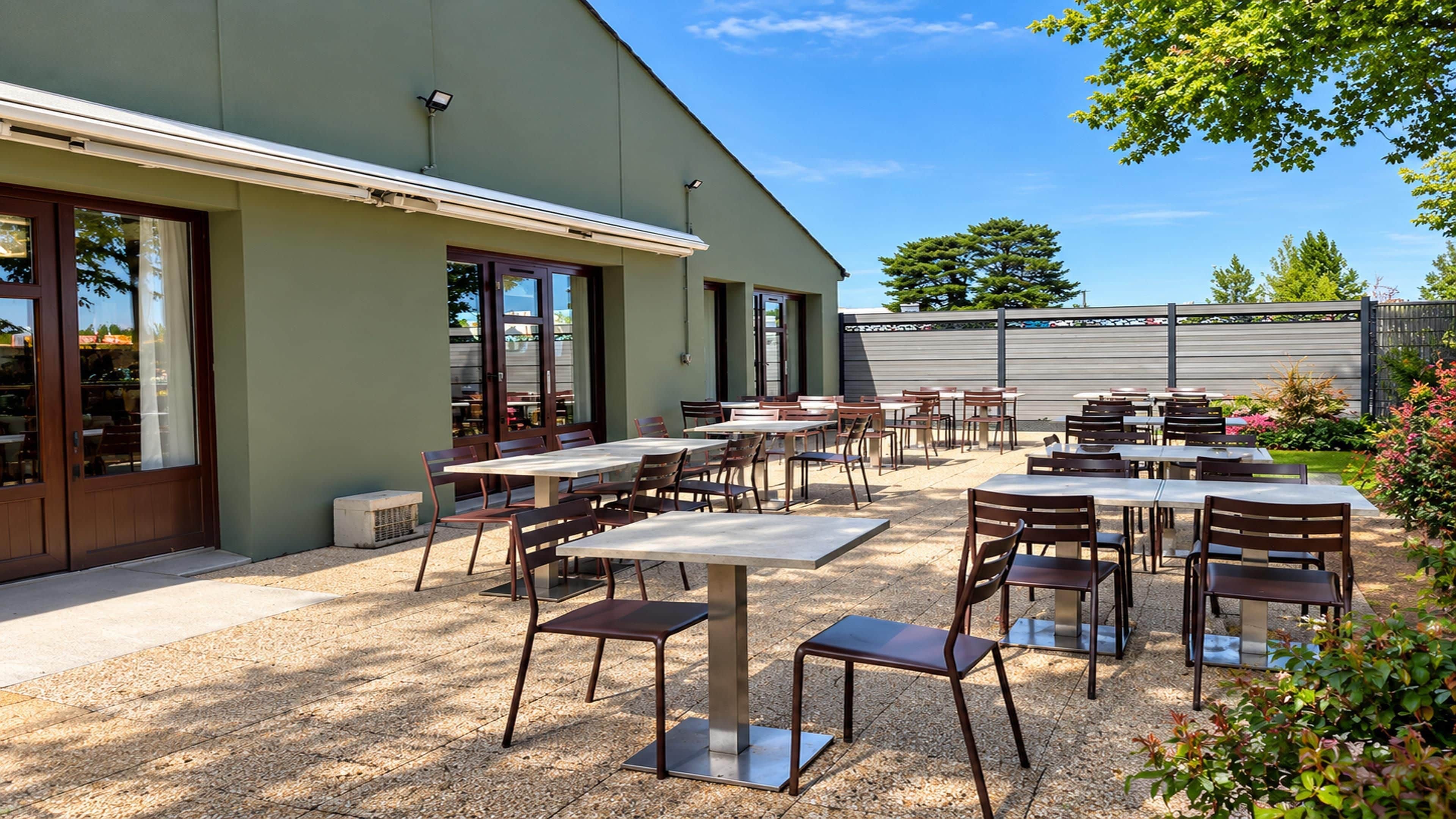 Une terrasse extérieure avec des tables et des chaises, un bâtiment doté de grandes fenêtres et un jardin clos.
