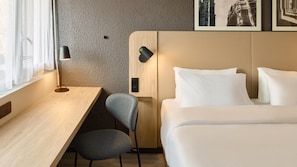 A hotel room with a wooden desk, a chair, a bed with white bedding, a wall-mounted lamp, and a framed picture. - Campanile NATURE - Nantes Ouest Saint Herblain (Saint-Herblain)