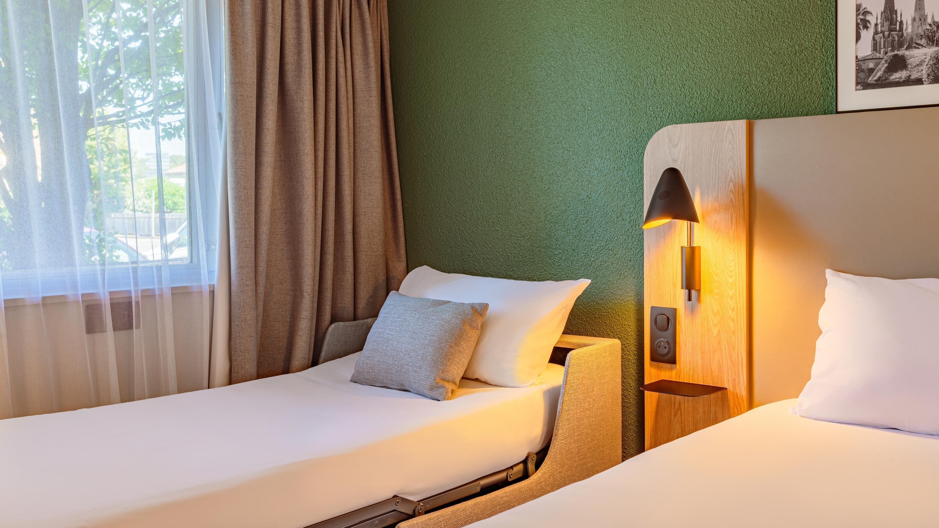 Une chambre d’hôtel avec deux lits, un mur texturé de couleur verte, une fenêtre avec des rideaux et une lampe fixée au mur.