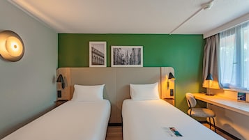 Quarto de hotel com duas camas, uma escrivaninha, uma cadeira, uma janela com cortinas e duas molduras na parede.