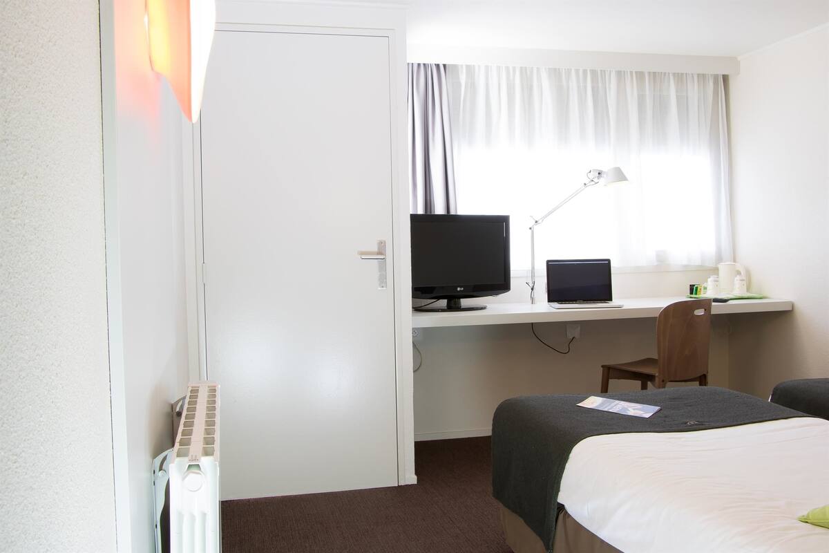 A hotel room with a bed, a desk with a computer, a chair, and a window with curtains.
