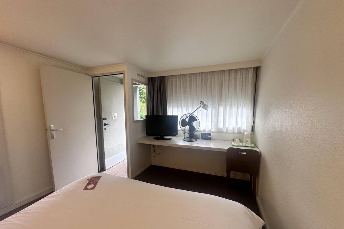 A hotel room with a bed, a desk with a computer, a TV, and a window with curtains.