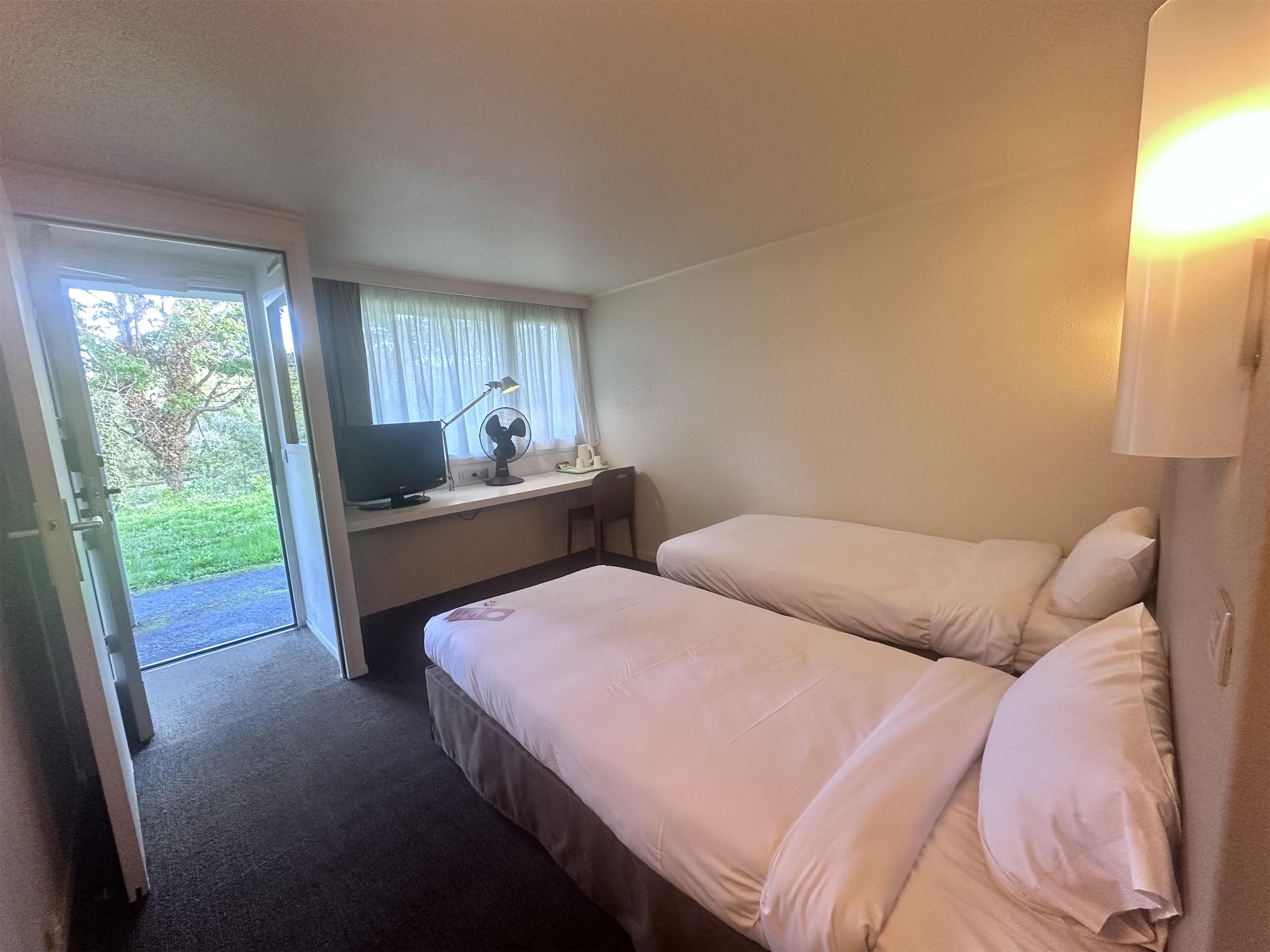 A hotel room with two beds, a desk with a computer and lamp, and a window with curtains.