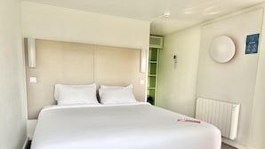 Un lit bien fait dans une pièce équipée d’un radiateur, d’une étagère verte et d’un luminaire fixé au mur.