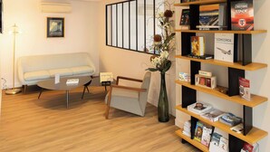 A modern hotel lobby with a sofa, a small table, a bookshelf with books and magazines, and a decorative vase with flowers. - Hotel Campanile Toulouse - Blagnac Aéroport (Blagnac)
