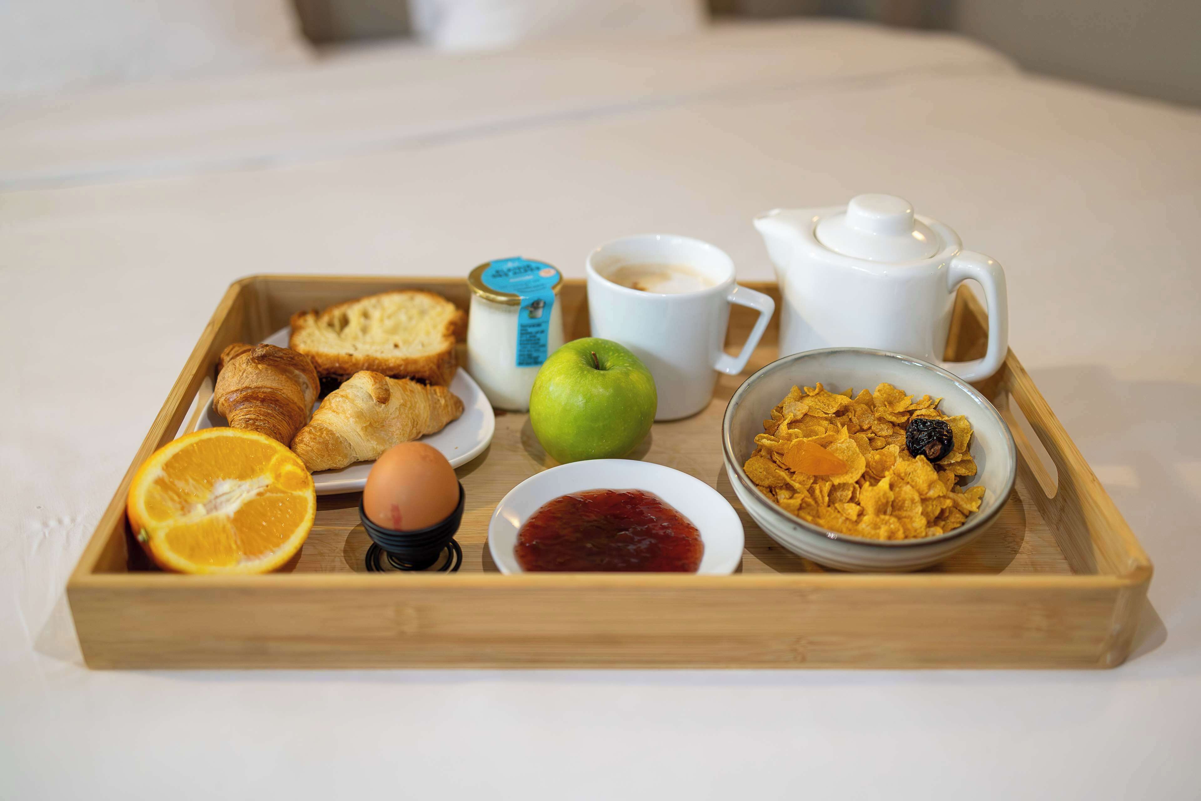 Un plateau de petit-déjeuner avec des croissants, une orange, une pomme, un œuf, de la confiture, des céréales, du café et une théière, posé sur un lit.