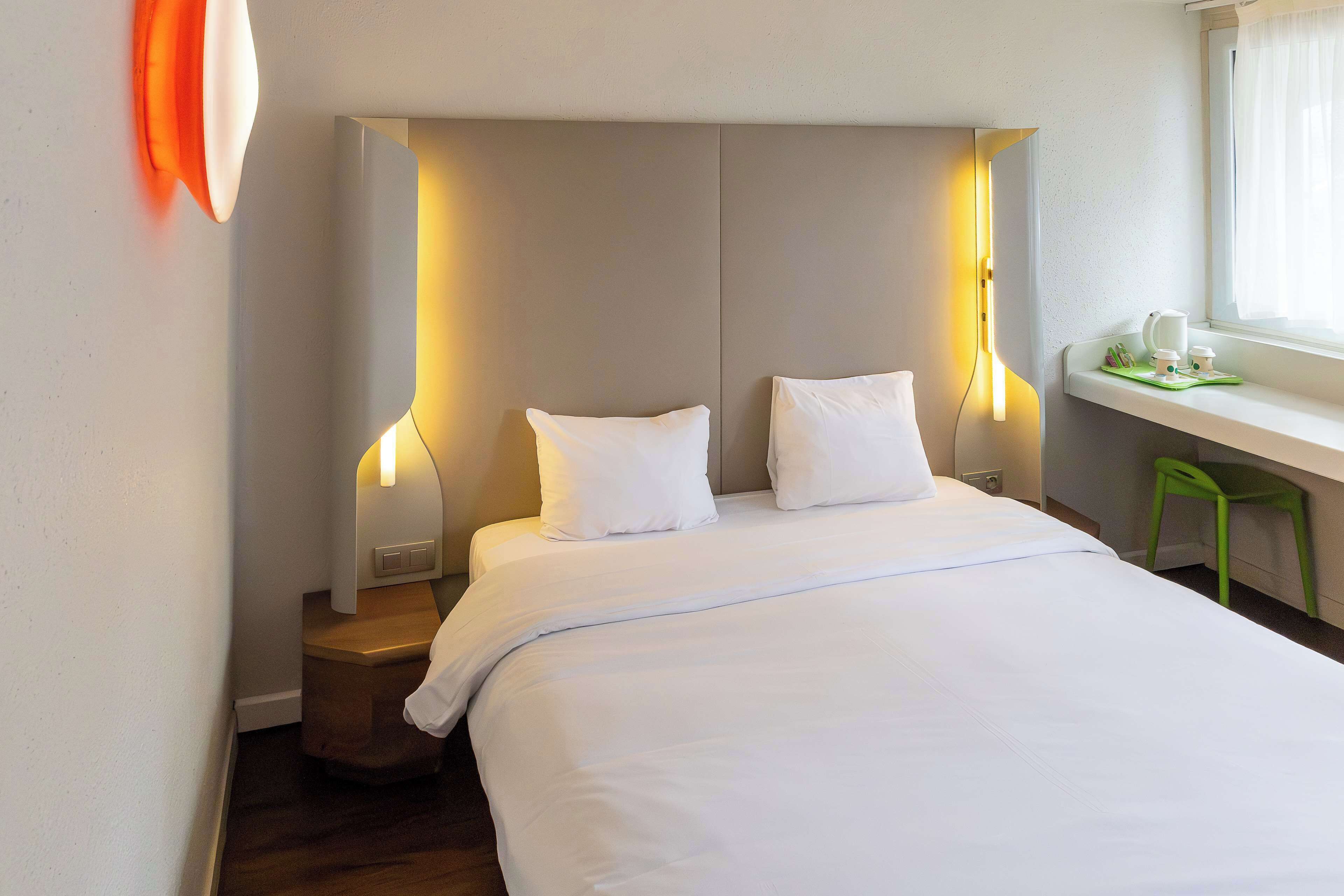 A neatly made bed with white linens and pillows, a bedside table with a lamp, and a window with curtains.