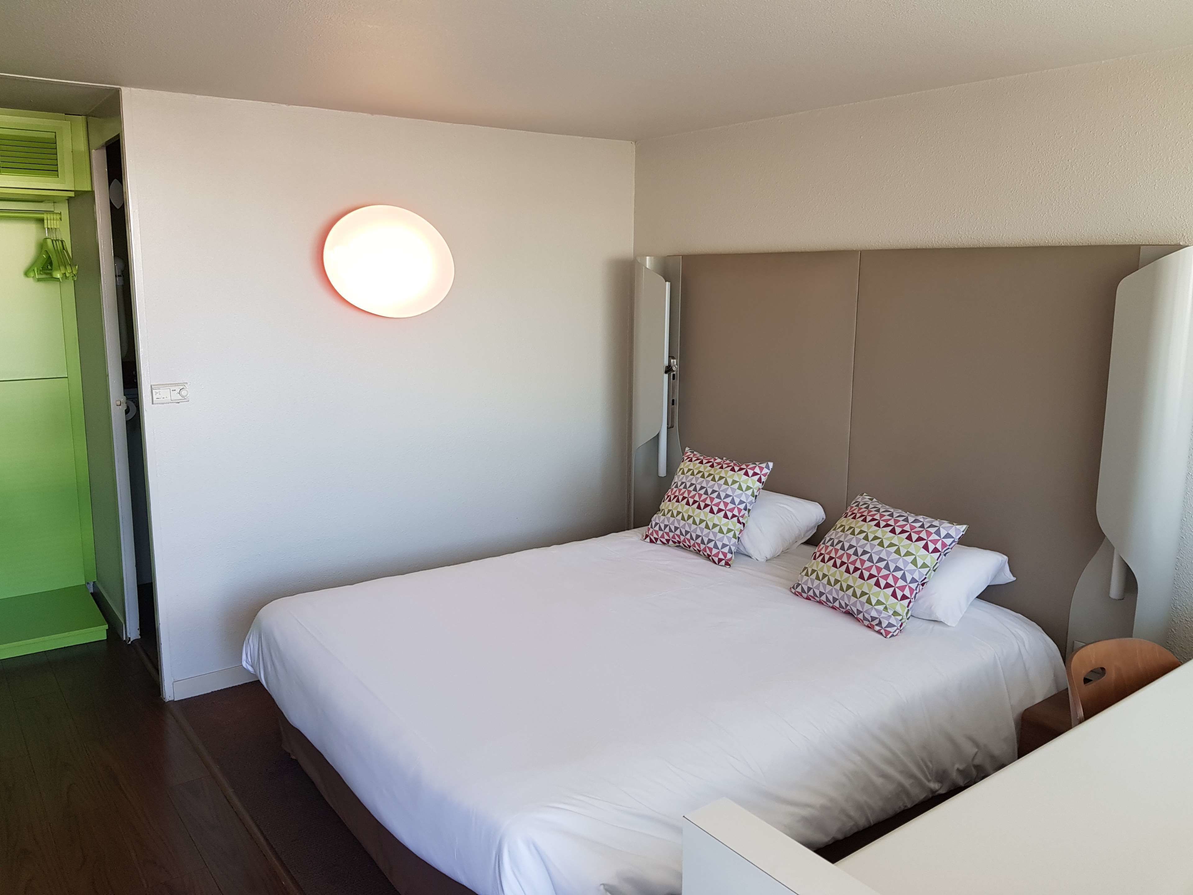 Una cama bien hecha con sábanas blancas y almohadas estampadas, una lámpara de pared redonda y un mueble verde en la esquina.