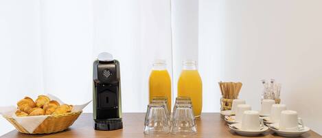 A breakfast setup with croissants, a coffee maker, orange juice, and white cups on a wooden table.