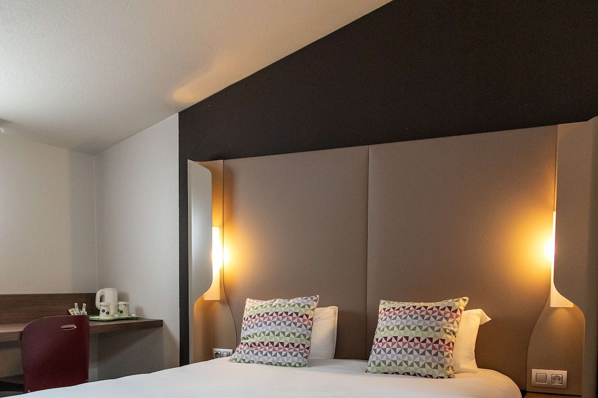 Quarto de hotel com uma cama grande, duas luminárias de cabeceira, uma escrivaninha com cadeira e um par de chinelos no chão.