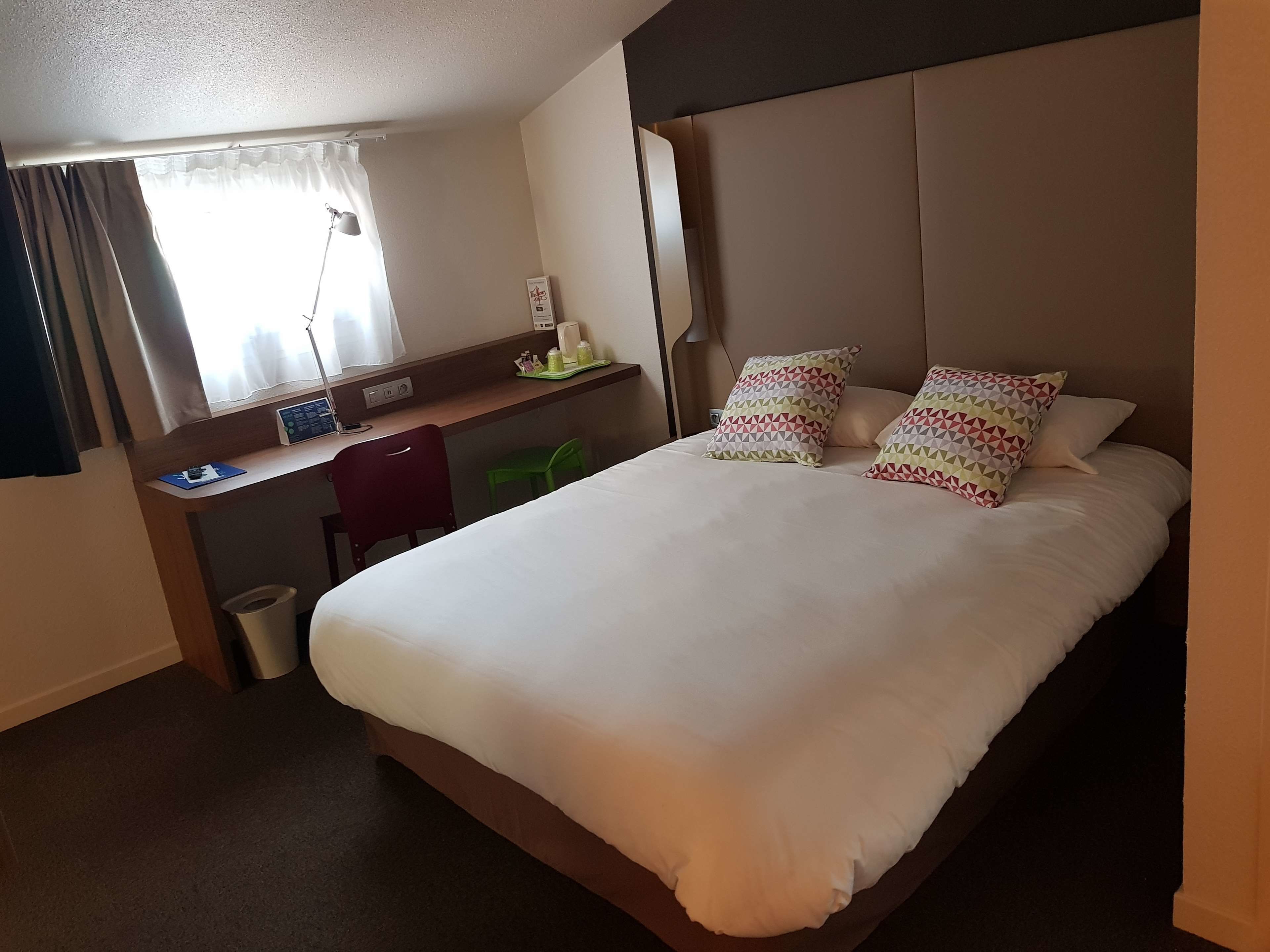 Habitación de hotel con una cama grande, un escritorio con silla, un cubo de basura pequeño y una ventana con cortinas.