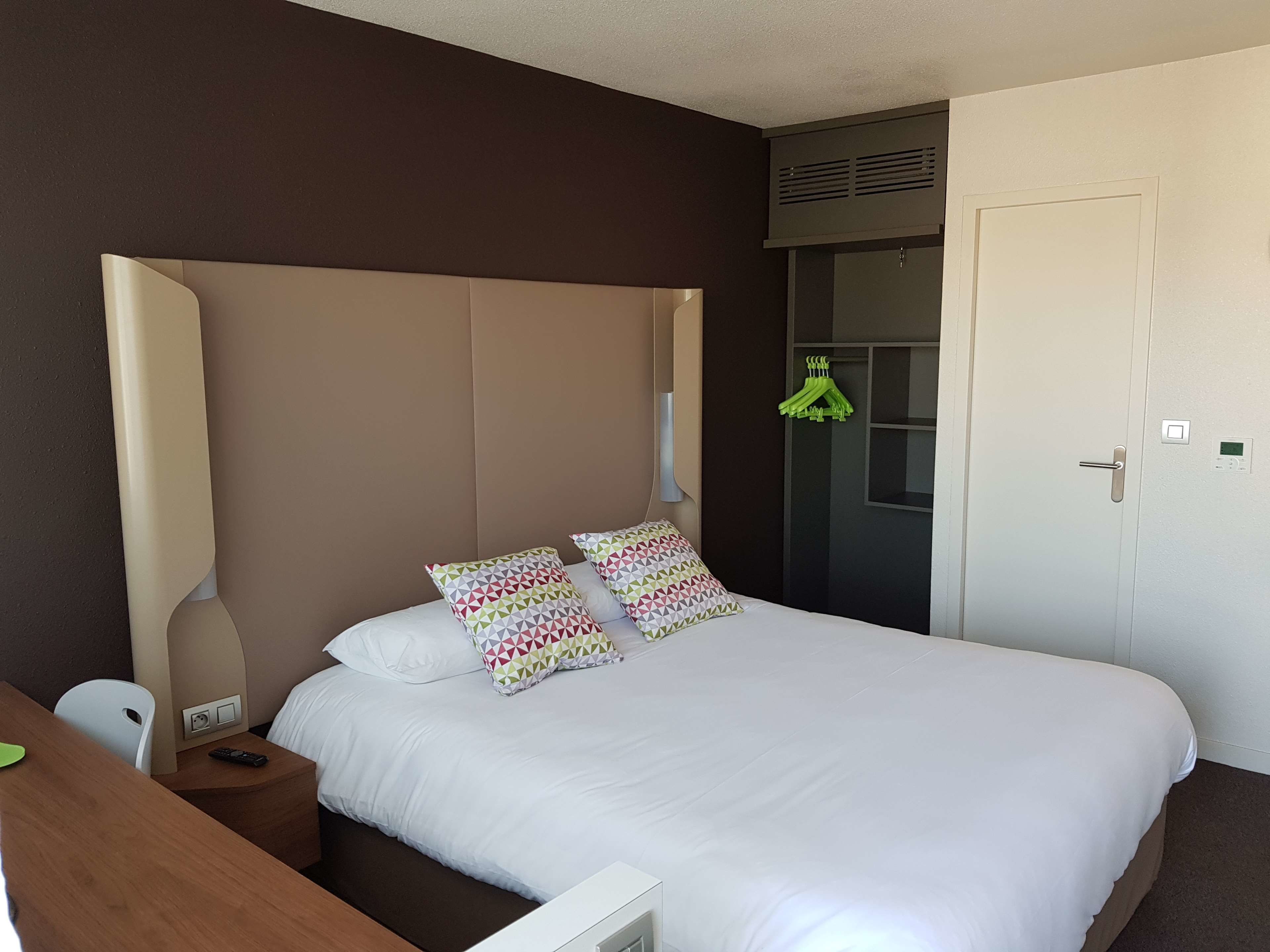 A hotel room with a large bed, two pillows, a bedside table with a remote control, a chair, and a wardrobe with a mirror and a hanging rack.