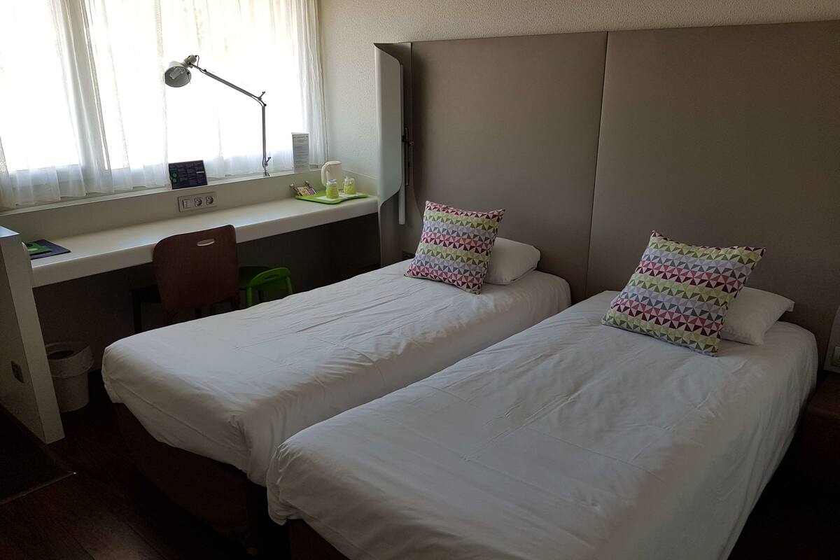 A hotel room with two single beds, a desk with a chair, a lamp, and a window with curtains.