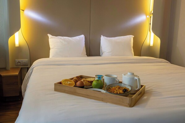 Una cama bien hecha con ropa blanca, una bandeja de desayuno con tostadas, fruta y una taza, y una mesita de noche con una lámpara.
