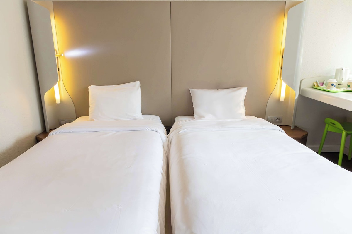 Quarto de hotel com duas camas de solteiro, uma mesinha de cabeceira com abajur e uma pequena escrivaninha com cadeira.