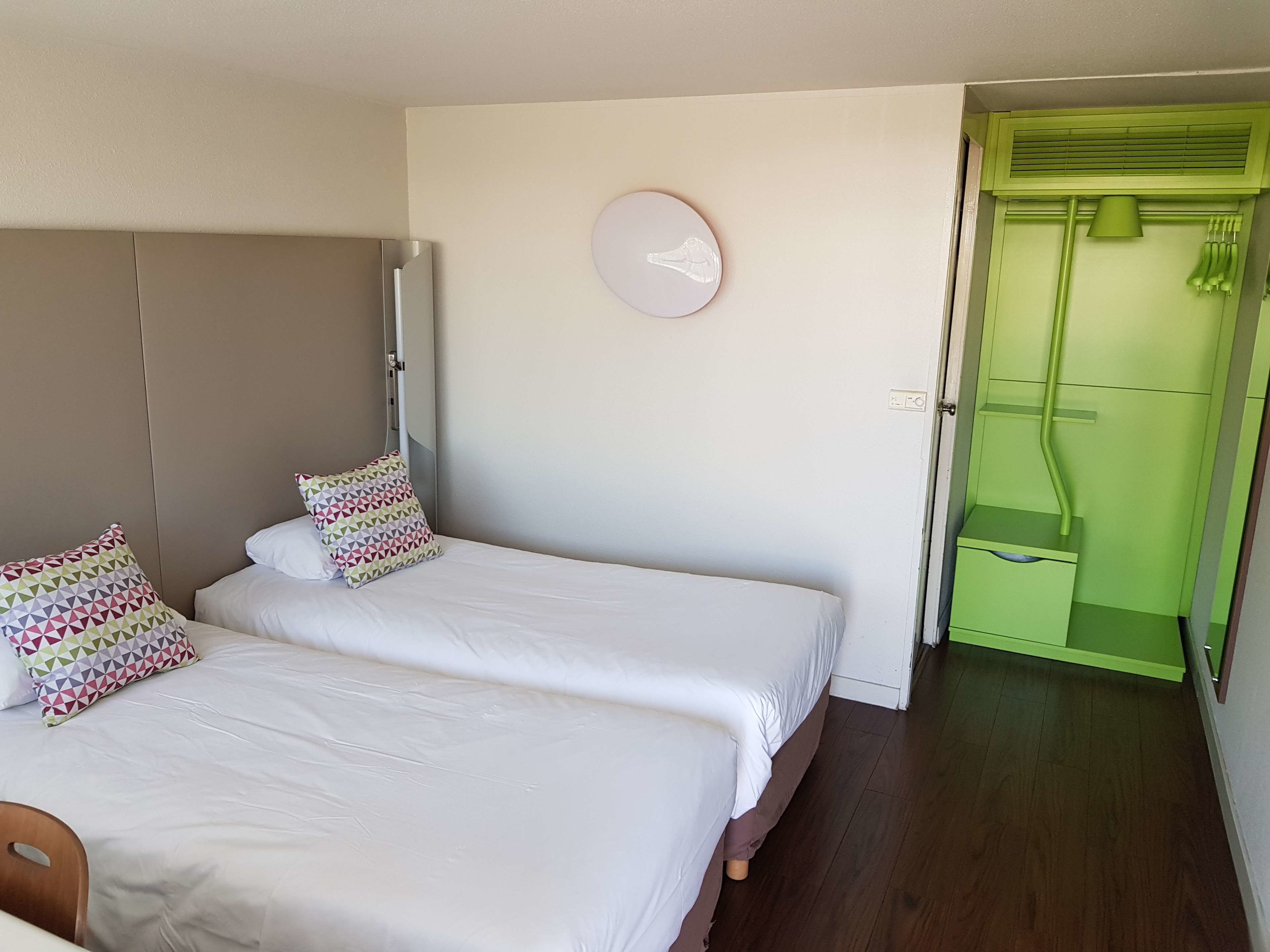 A hotel room with two beds, a wardrobe, a green bathroom, and a wall clock.