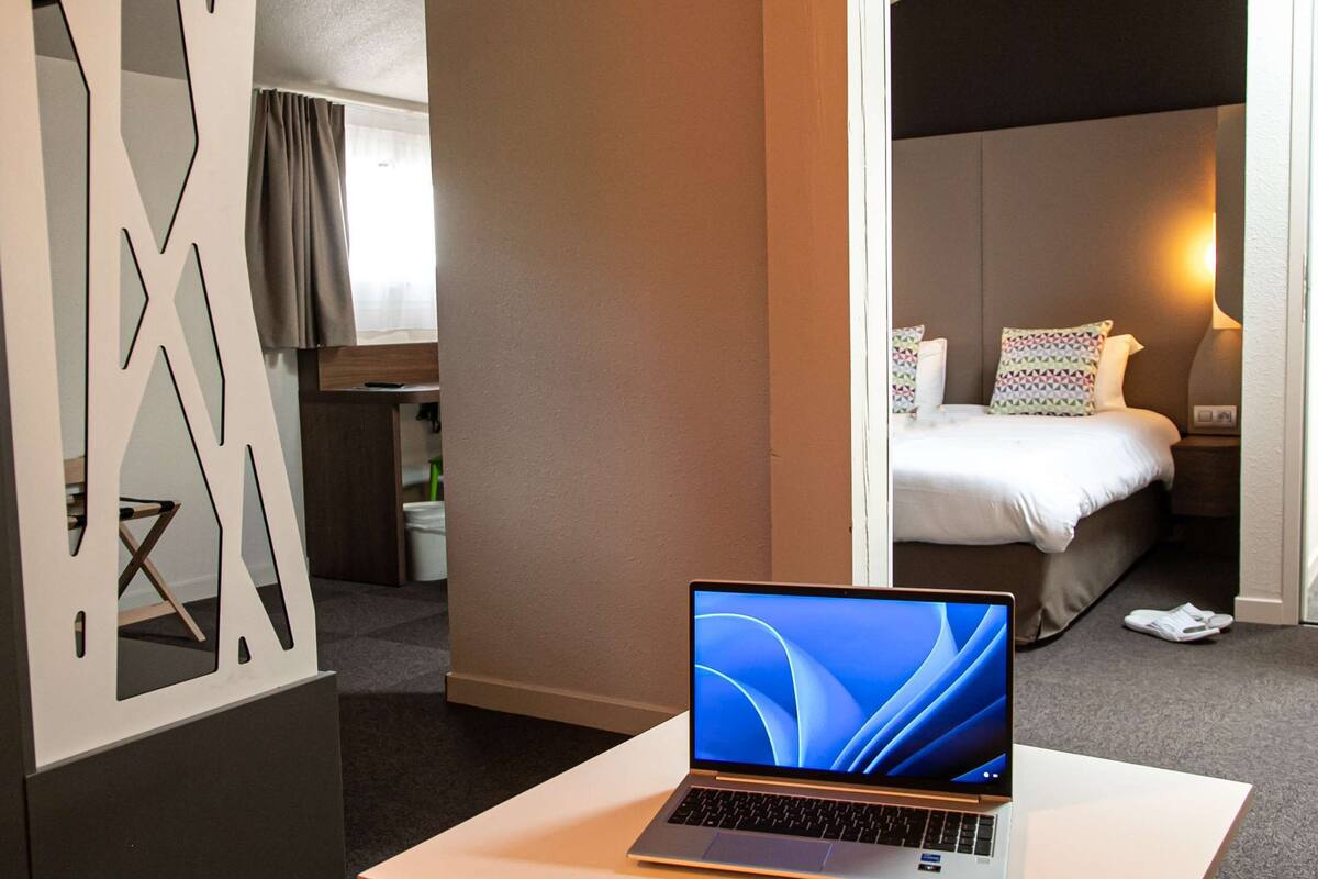 A hotel room with a laptop on a table, a bed with pillows, a nightstand, and a window with curtains.
