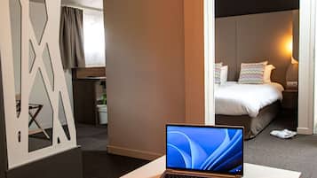 Una habitación de hotel con una computadora portátil sobre una mesa, una cama con almohadas, una mesita de noche y un ventanal con cortinas.