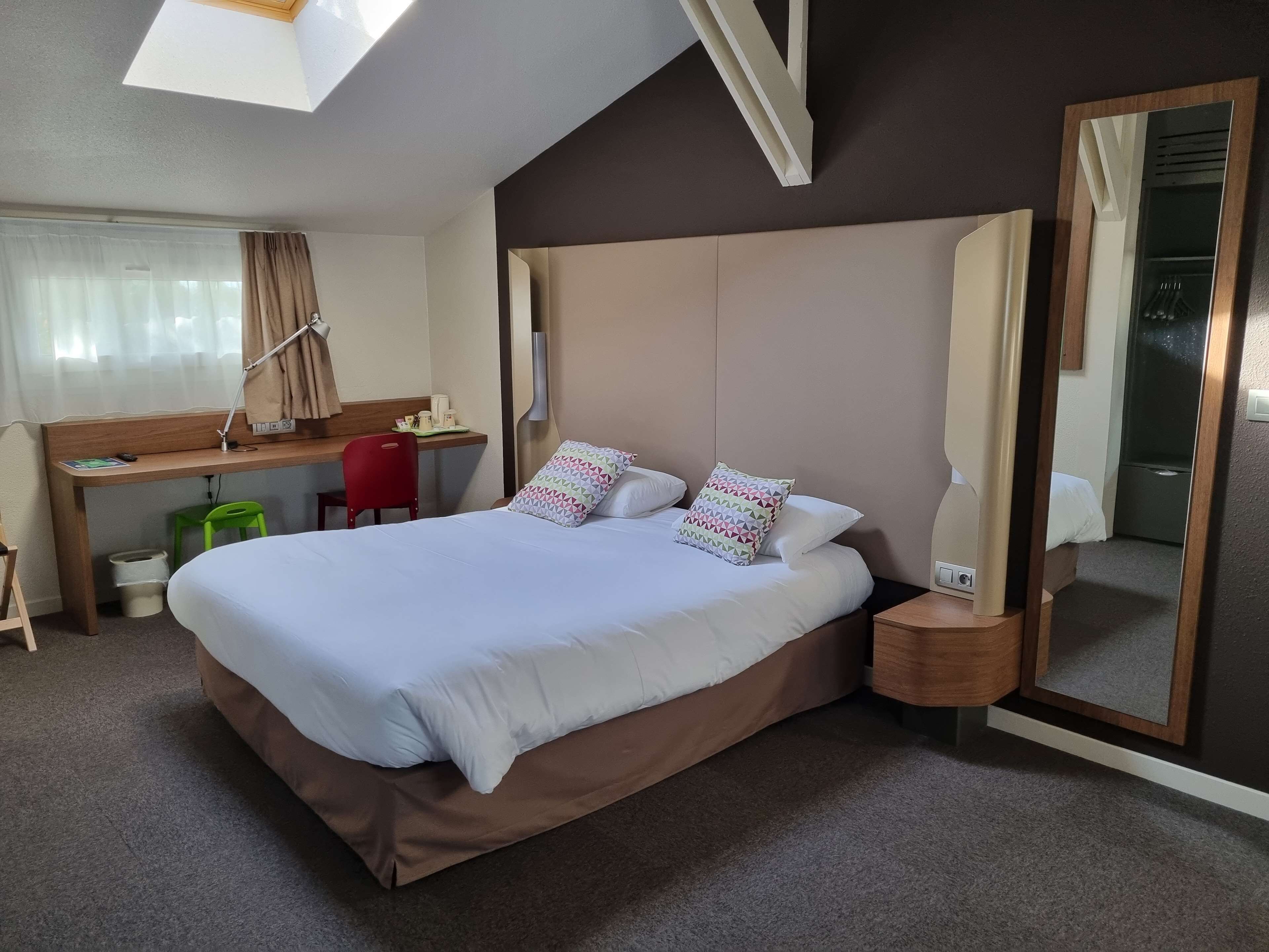 Una habitación de hotel con una cama grande, un escritorio, una silla, un espejo y un tragaluz.