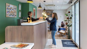 A reception area with a wooden reception desk, a bulletin board with photos, and a seating area with a plant and a chair. - Hotel Campanile Toulouse - Blagnac Aéroport (Blagnac)