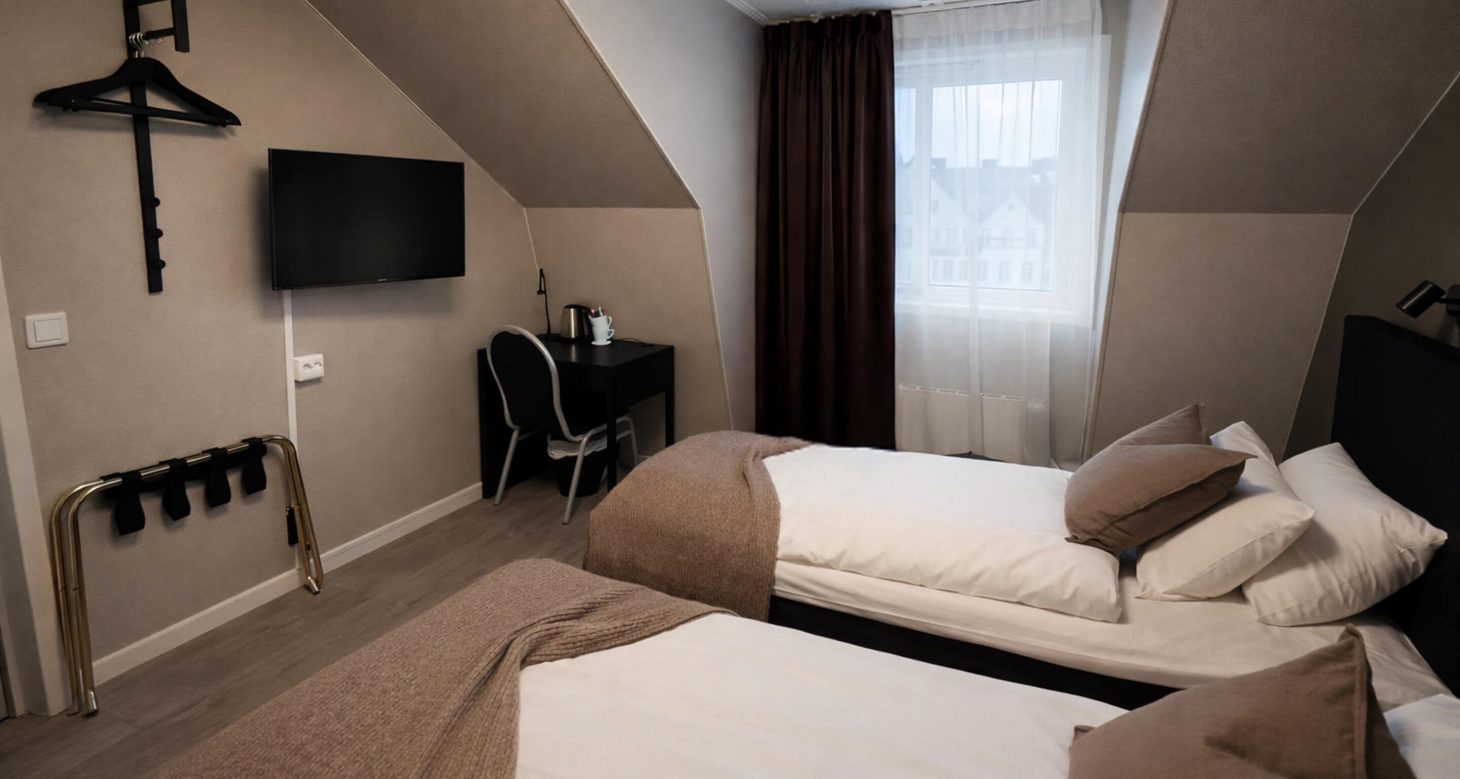 Een hotelkamer met twee bedden, een bureau met stoel, een aan de muur bevestigde televisie en een raam met gordijnen.