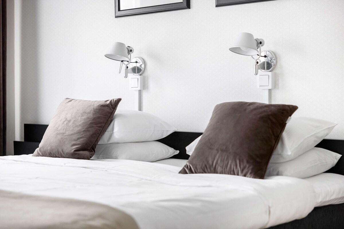 A neatly made bed with white and brown pillows, two wall-mounted lamps, and a framed picture above the bed.