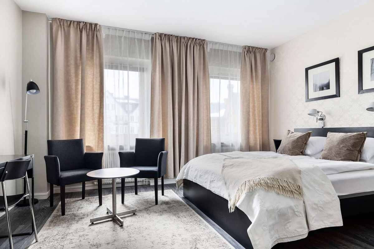 Moderni hotellihuone, jossa on suuri sänky, kaksi nojatuolia, pieni pyöreä pöytä ja lattiavalaisin.