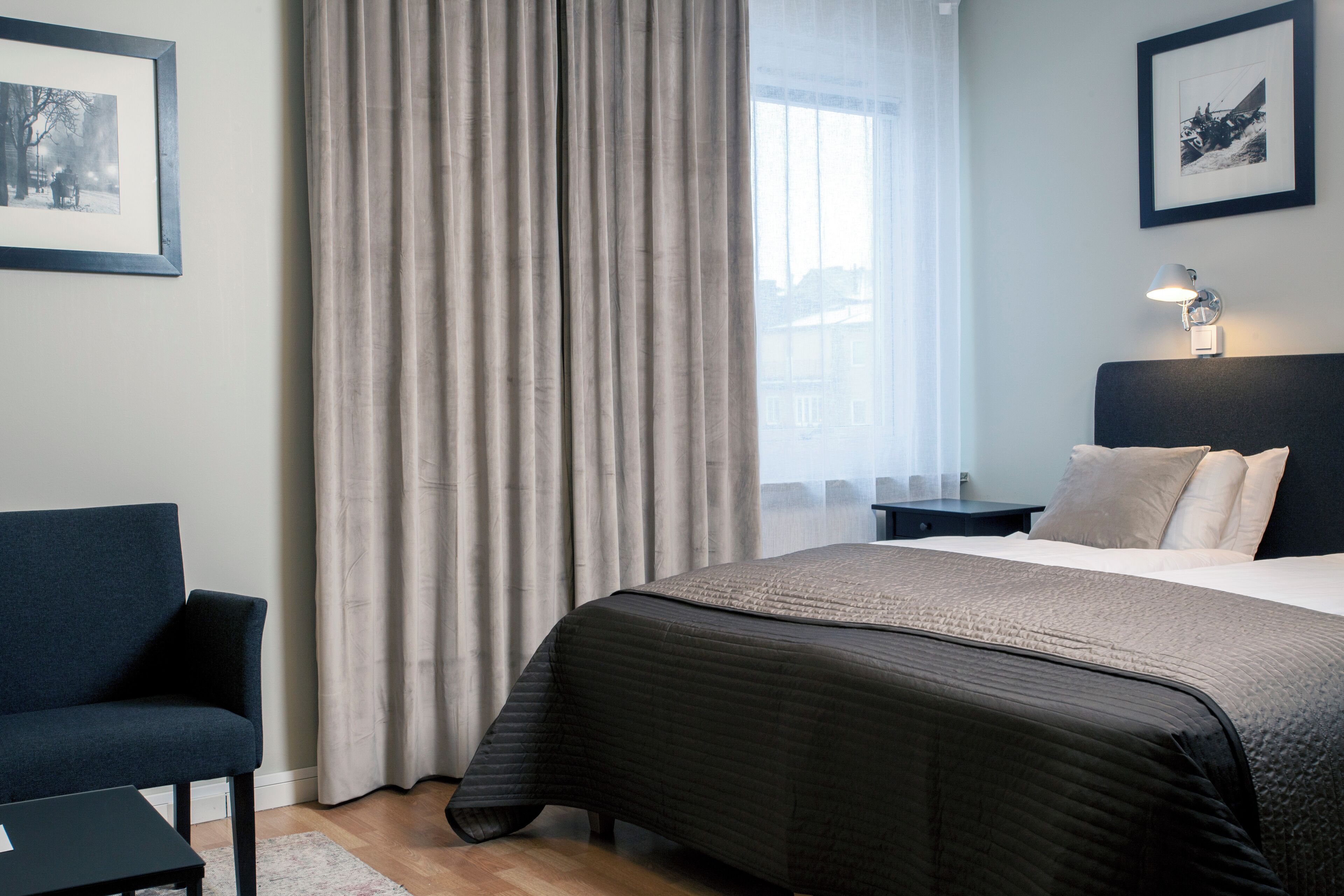 Une chambre d’hôtel moderne avec un grand lit, un fauteuil sombre, une table de chevet et une fenêtre avec des rideaux.