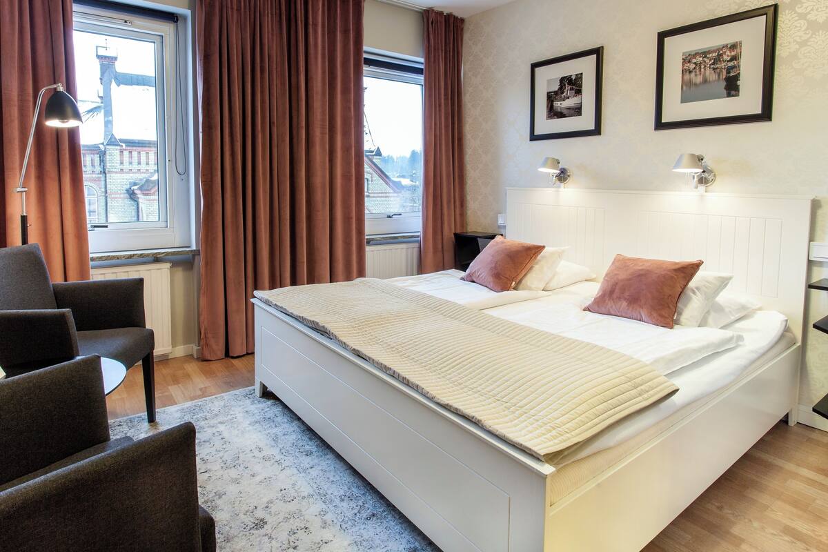 Ein Hotelzimmer mit einem großen Bett, zwei Nachttischen, einem Sessel, einem Schreibtisch und Blick auf ein Gebäude durch ein Fenster.