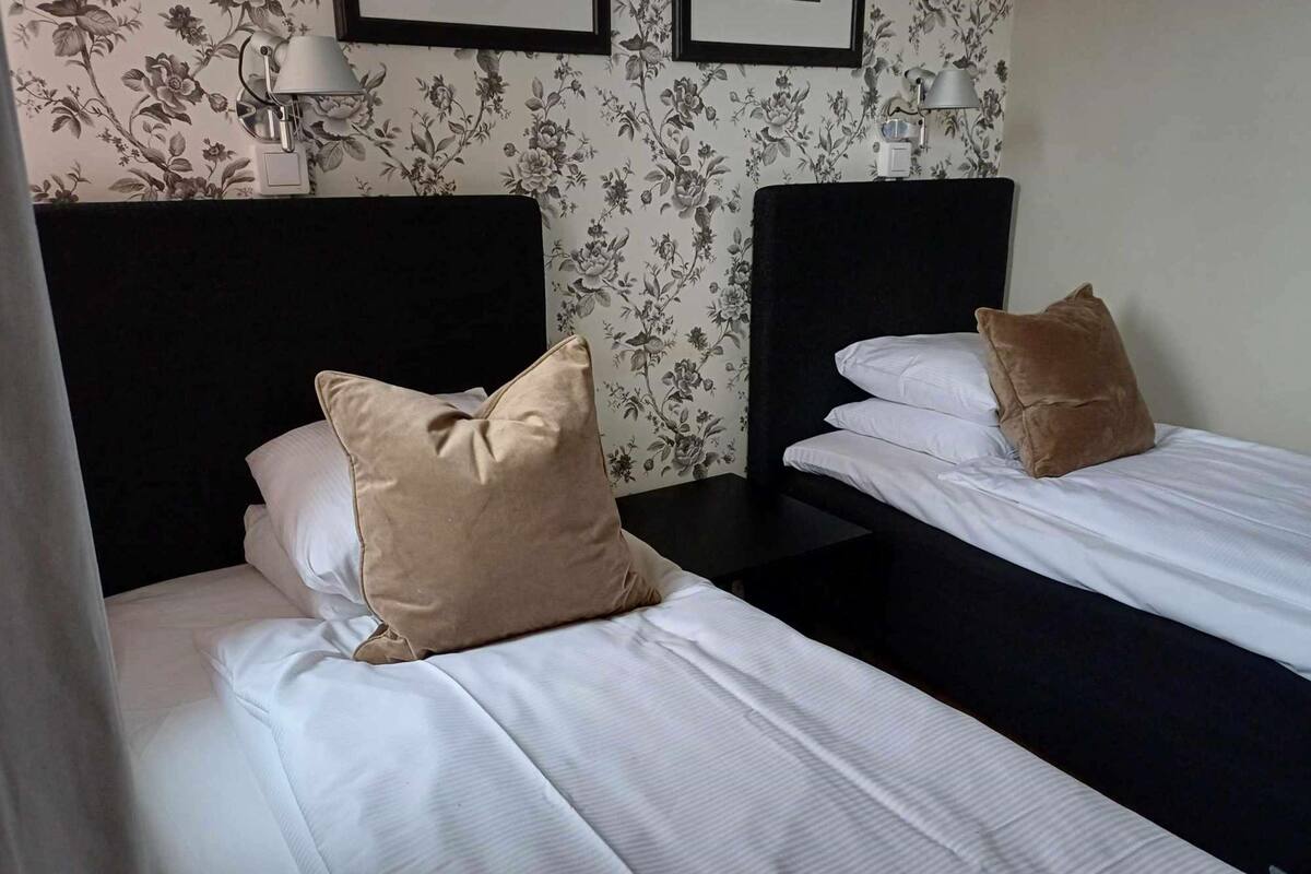 Zwei Einzelbetten mit weißen Leinen und beigen Kissen, ein schwarzes Kopfstück und ein Wandpapier mit floralem Muster.