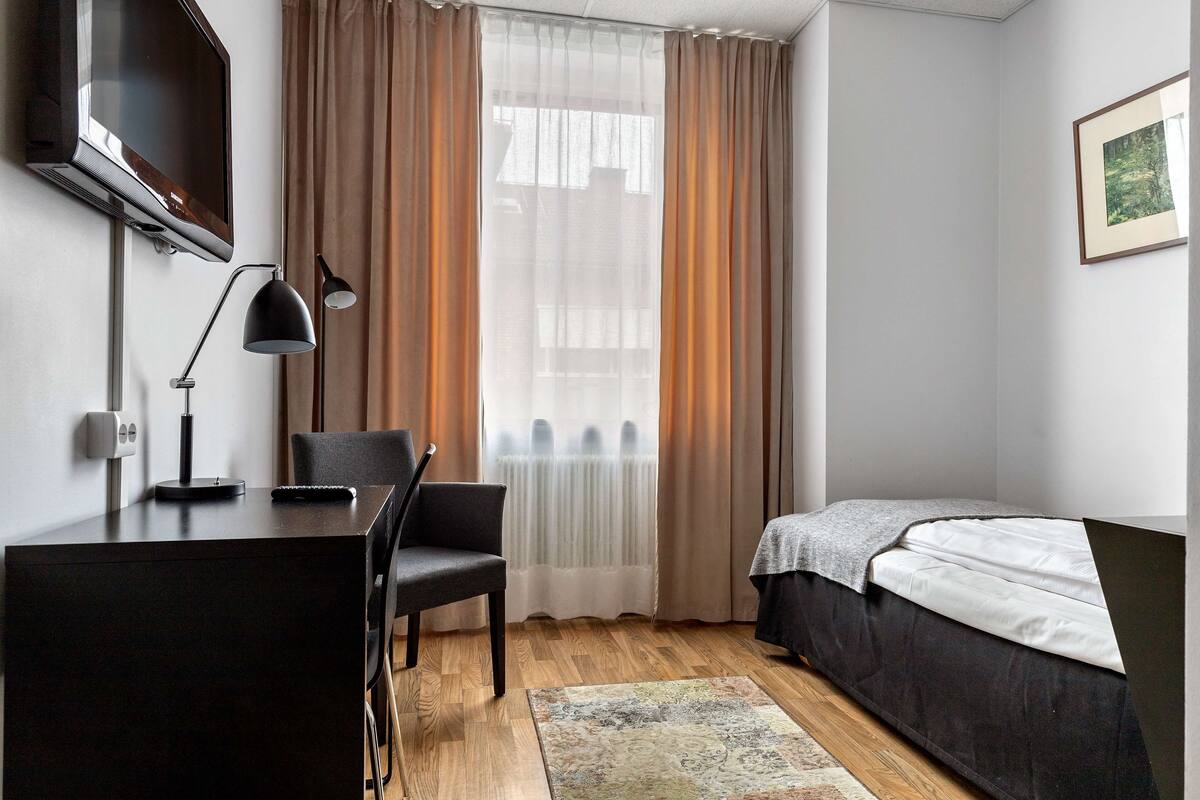 Ein Schlafzimmer mit Bett, Schreibtisch mit Stuhl, Fernseher und einem Fenster mit Vorhängen.