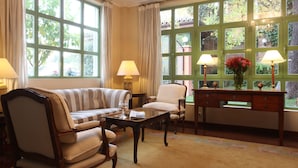 A cozy living room with a sofa, armchairs, a coffee table, and a desk with lamps. Large windows with curtains allow natural light and views of greenery outside. - Hotel Quinta de los Cedros (Madrid)