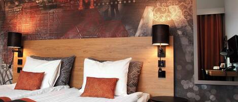 A hotel room with a large bed, patterned pillows, a headboard with wall art, two wall-mounted lamps, and a small table.