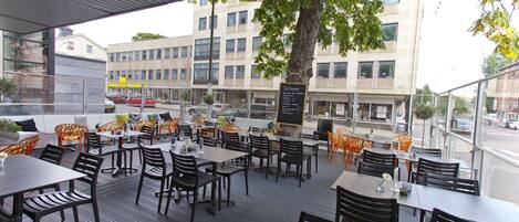 An outdoor dining area with tables and chairs, a menu board, and a view of the street and buildings in the background.