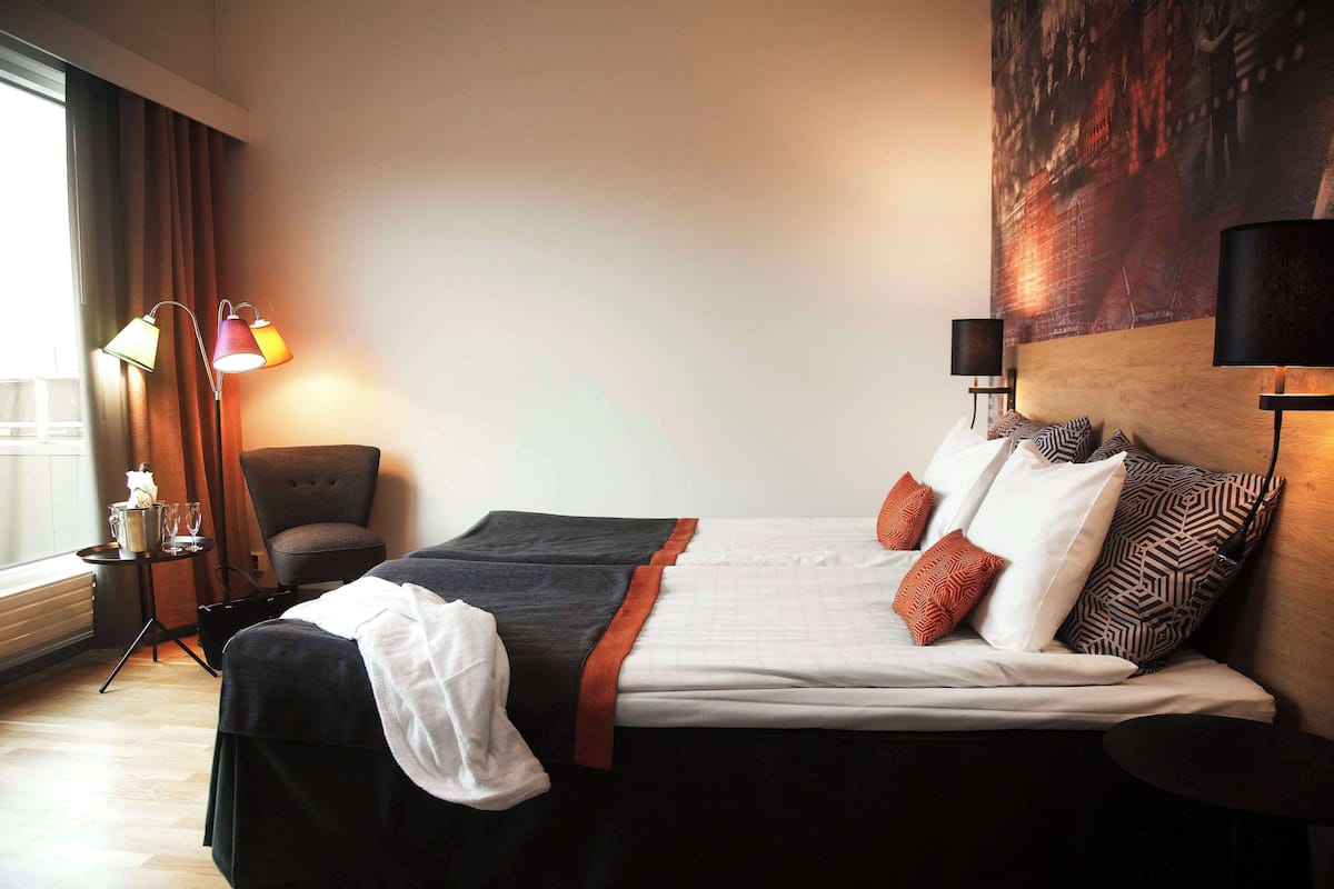 Ein Hotelzimmer mit einem Bett, zwei Nachttischlampen, einem Sessel, einem kleinen Tisch und einem großen Fenster mit Vorhängen.